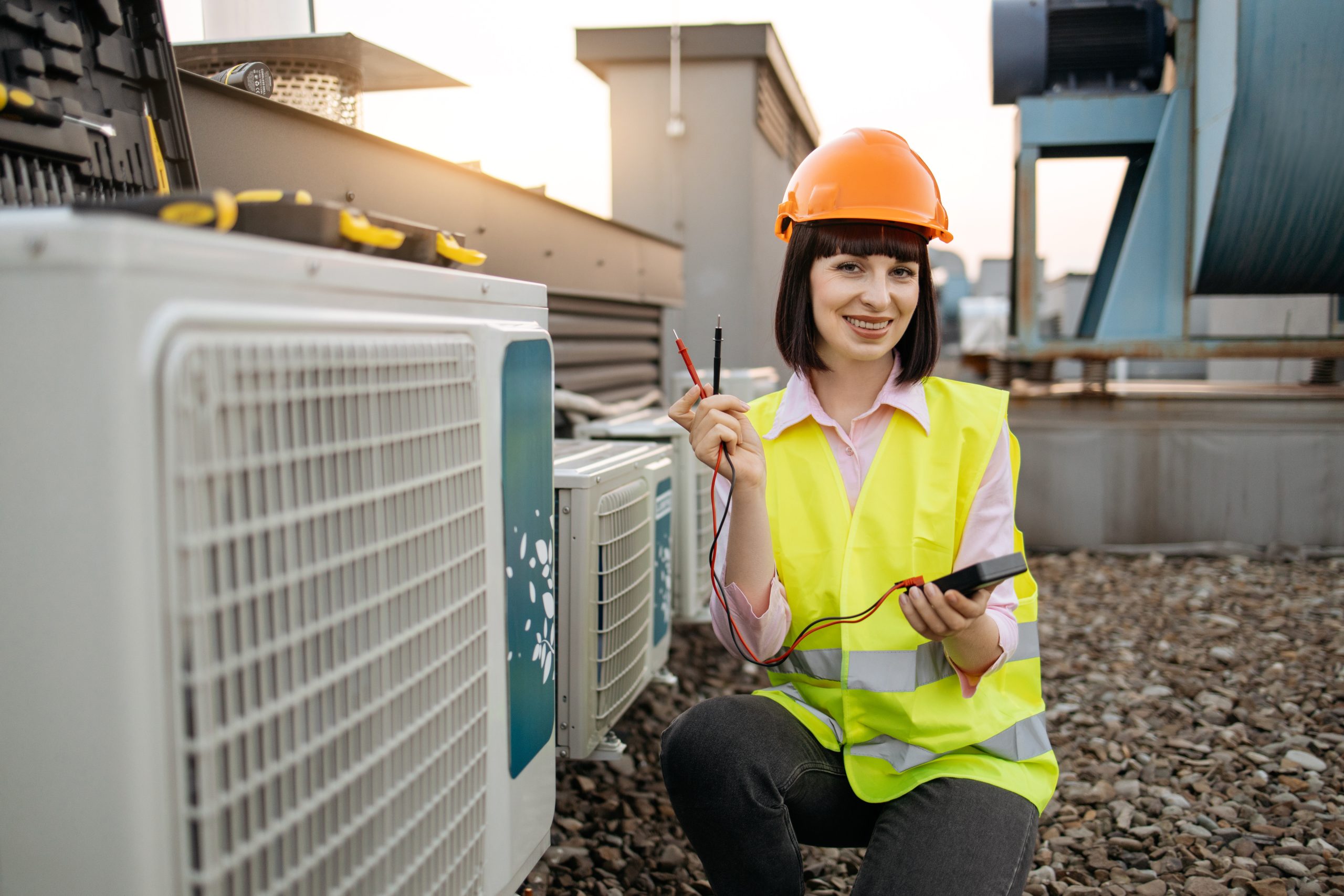 Positive caucasian female getting ready to start measuring voltage with digital multimeter while sitting next to large cooling system on rooftop. Concept of repairing and air conditioner.